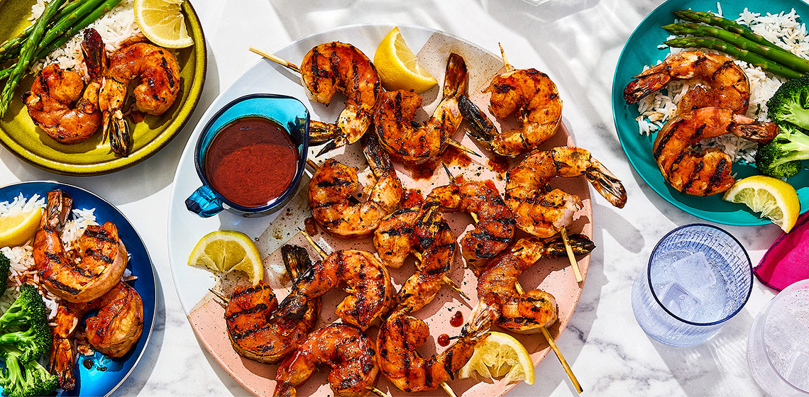 Overhead shot of four plates of Korean-style fiery shrimp skewers, with accompanying sides including rice, broccoli and lemon wedges.