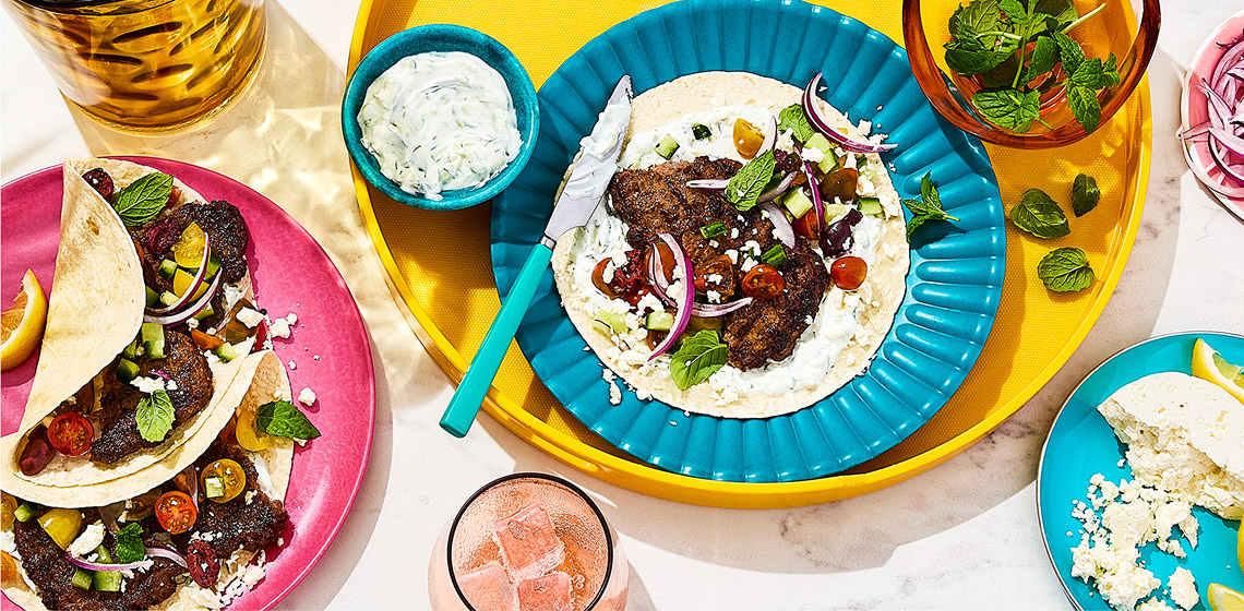 Overhead shot of four Greek-inspired smashed burger tacos with tzatziki on the side.