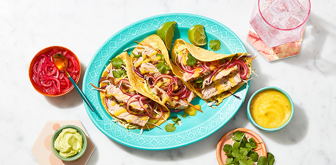 A teal plate with three grilled mahi mahi tacos and lime wedges with small bowls with toppings all around it.