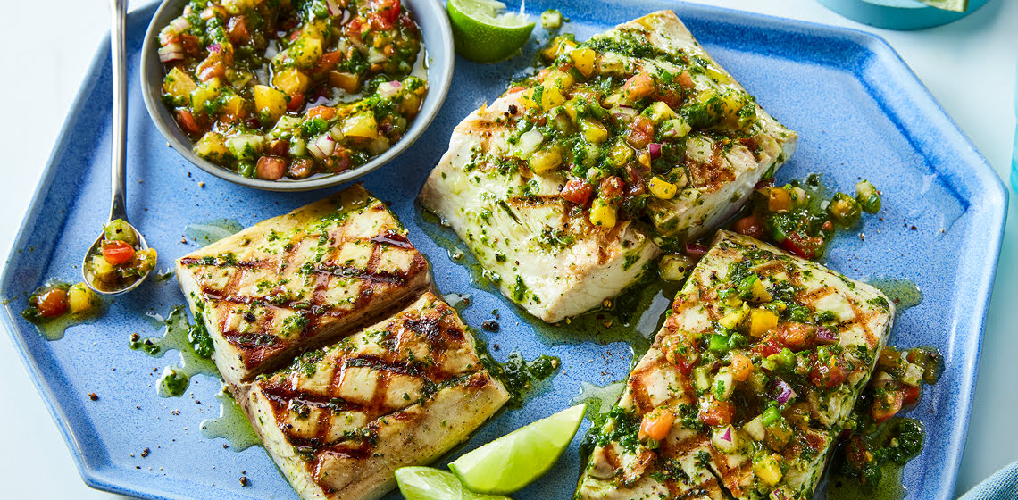 Three pieces of grilled mahi mahi with a green sauce, a bowl of just the sauce, a spoon with some sauce on it and lime wedges.