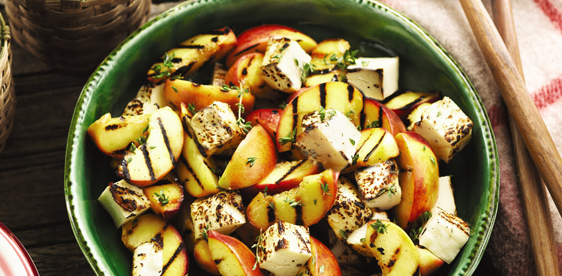 A green bowl filled with grilled peach and haloumi salad on a picnic bench with wooden serving spoons.