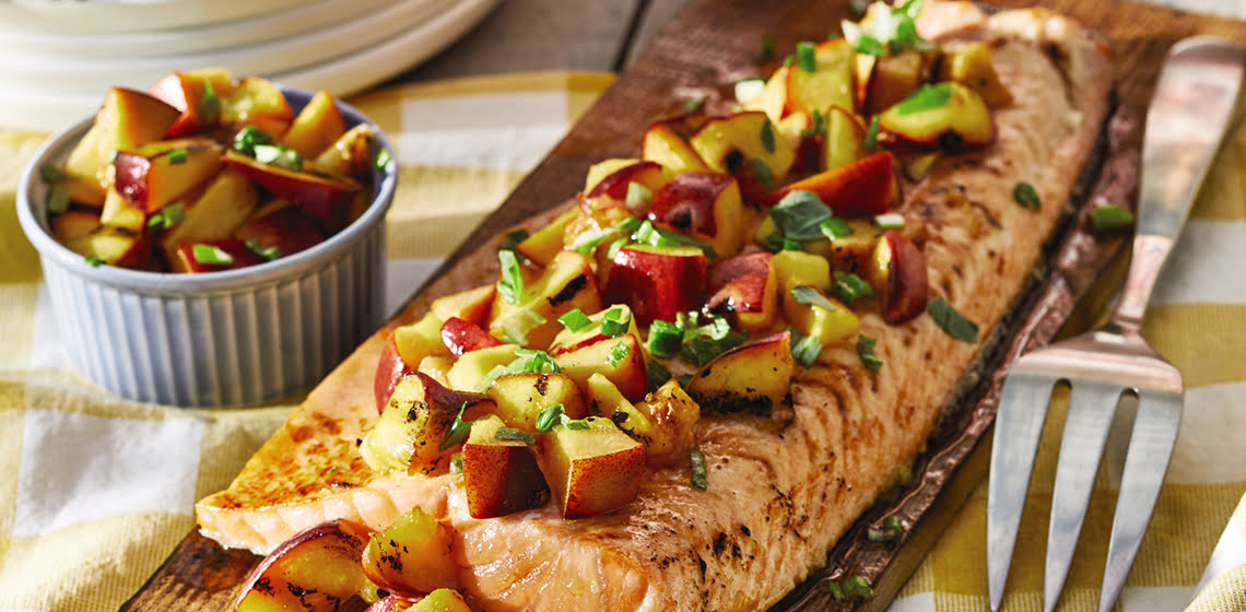 Grilled salmon on a plank with grilled peaches on top, a serving fork beside it, and a small bowl of just the peaches off to the other side.