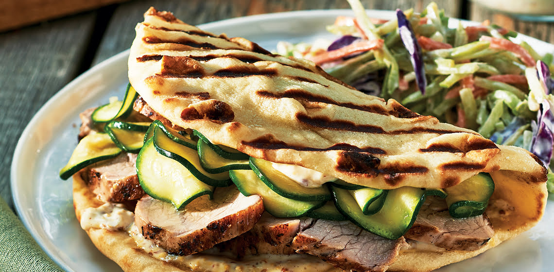 A pork and zucchini pickle sandwich in naan with a slaw on a white plate.