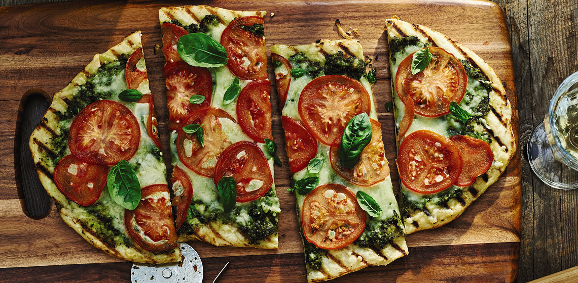 A cut up grilled tomato pizza on a wooden board with a pizza cutter also on the board and some filled glasses around it.