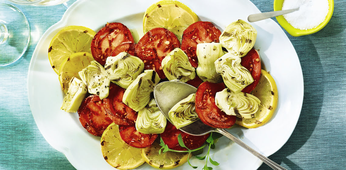 Grilled tomatoes, artichokes and lemons on a white plate with a spoon, and two wine glasses with white wine on a blue placemat