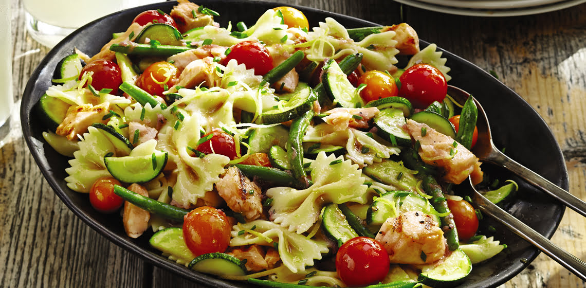 Large black bowl with grill vegetable and salmon bow tie pasta with two serving spoons.