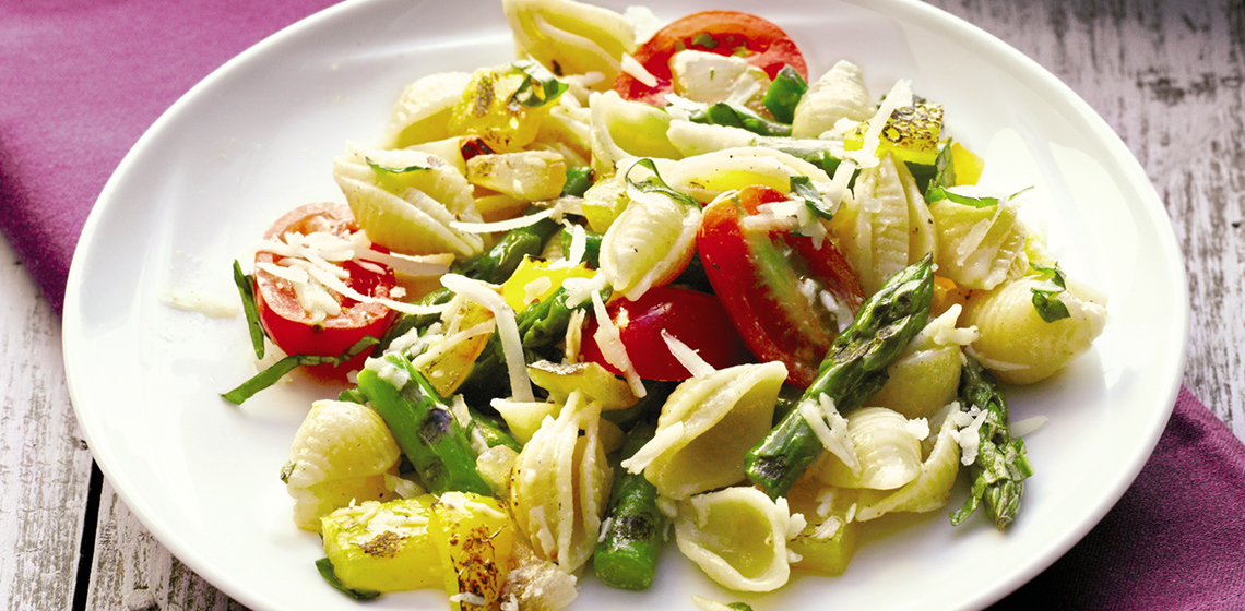 White plate with a pasta salad on a purple napkin and a large platter with more salad and serving spoons in the background.