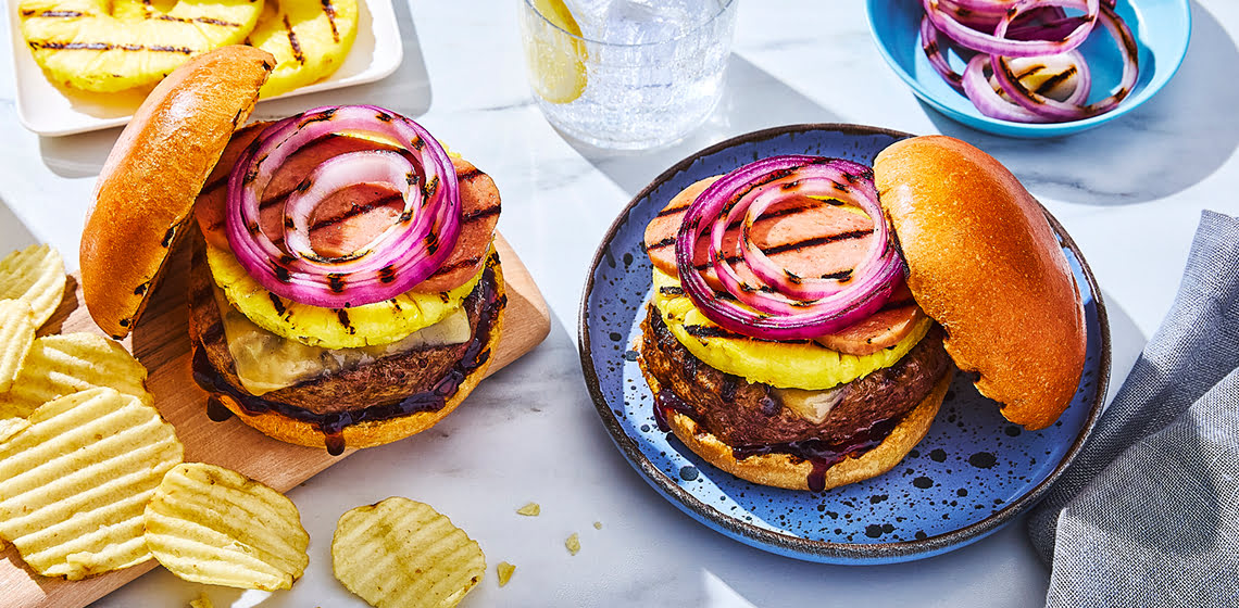 Two Hawaiian-inspired burgers on a white background with grilled pineapples and chips and two glasses of lemon water.