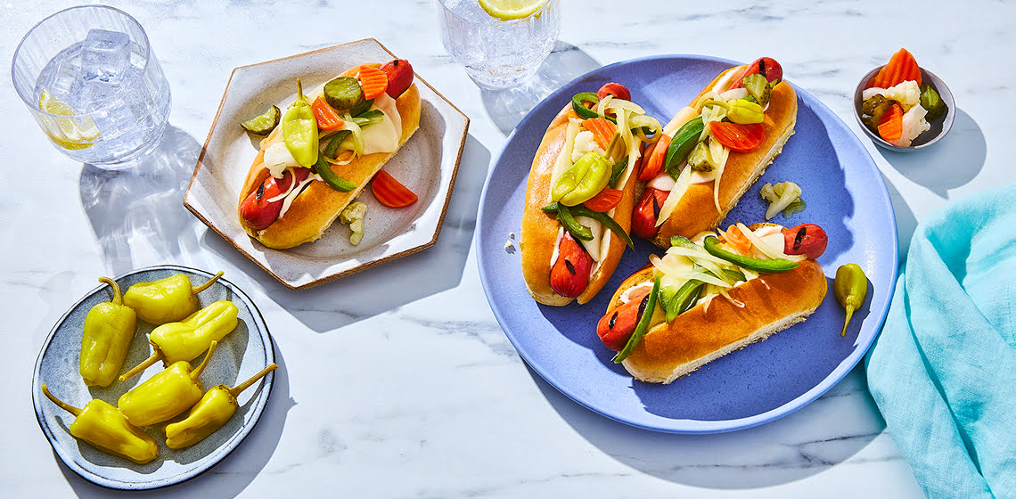 Italian-style hot dogs on a blue plate with toppings and a plate of hot peppers to the side.