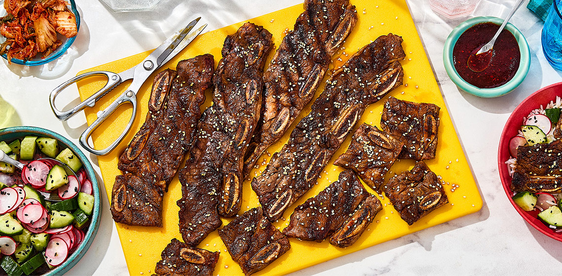 Overhead shot of kalbi Korean-style ribs on a yellow platter with scissors and a bowl of fresh salad on the side.