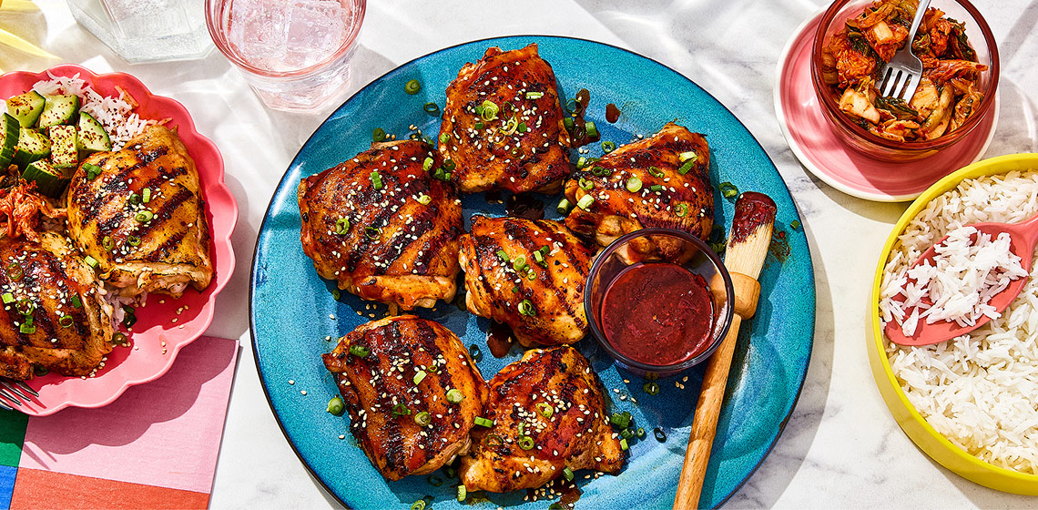 Two plates of Korean-style BBQ chicken, a bowl of kimchi and a bowl of rice on a marbled surface.