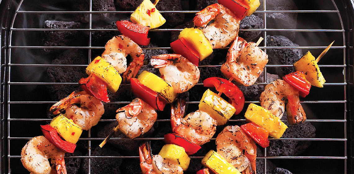 A charcoal grill with four shrimp kabobs with red pepper and pineapple.