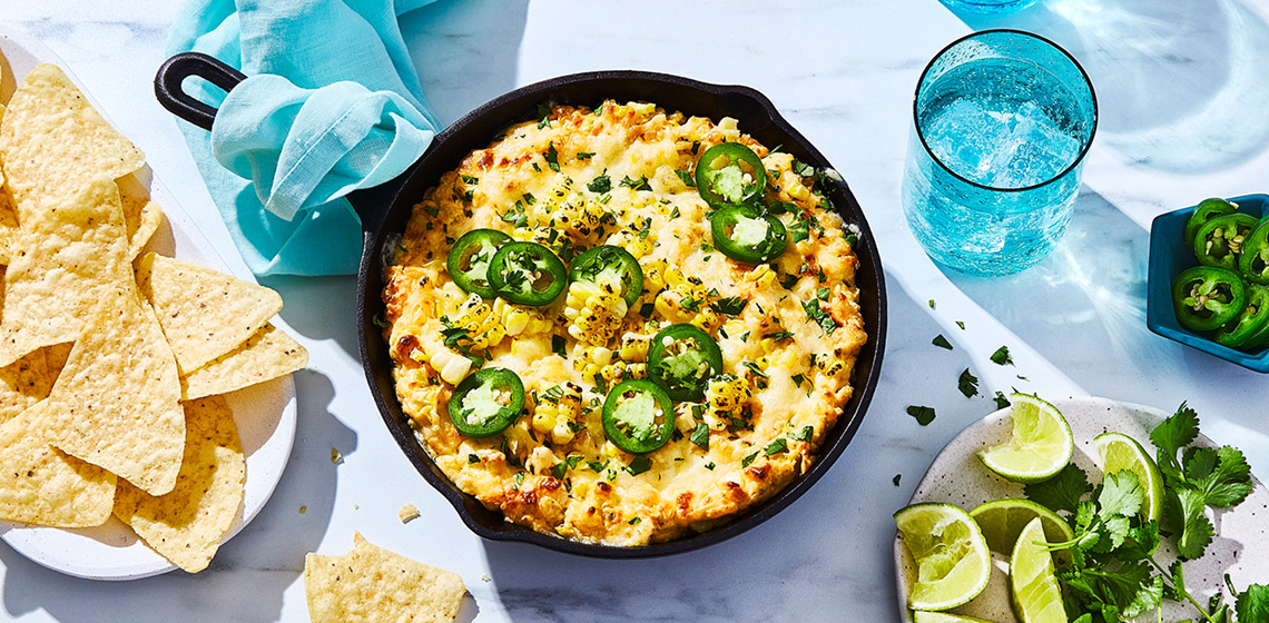 Overhead shot of Mexican-inspired Street Corn Party Dip in a skillet next to a plate of tortilla chips on a white plate, and cut limes and cilantro on a side dish.