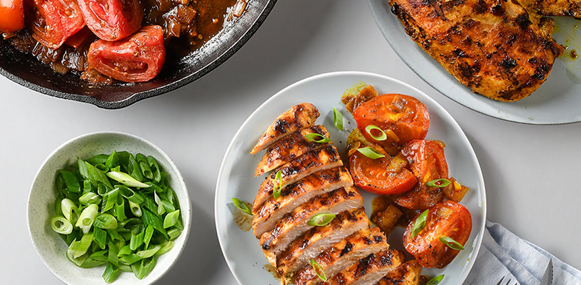 A plate of Mumbai style chicken with tomatoes, a small bowl of green onion, another plate with just chicken and another pan with the roasted tomatoes.