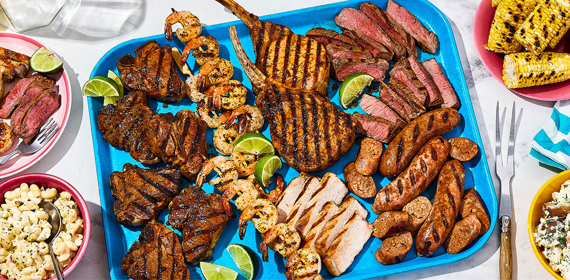 A large platter of all the Braai meats and seafood with a small plate of some off to the side, a small plate of grilled corn and two bowls with a pasta and potatoes.