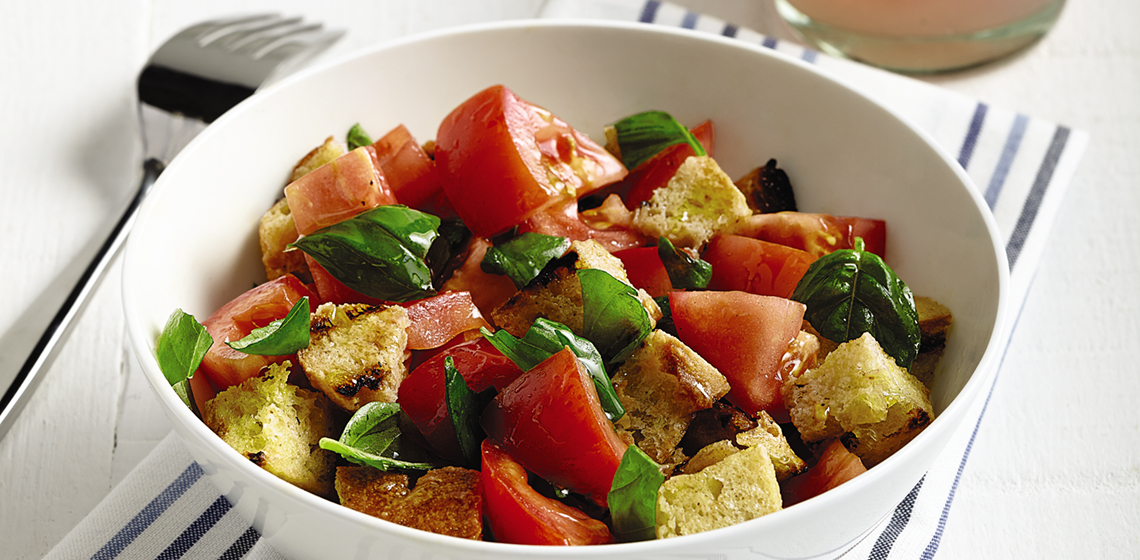 Bowl filled with grilled bread and tomato salad on a napkin with a fork beside it and a drink with lemon and ice.