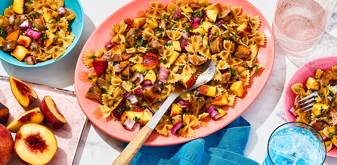 Platter filled with bowtie pasta salad and fresh peaches, with two bowls with the same pasta to the side.