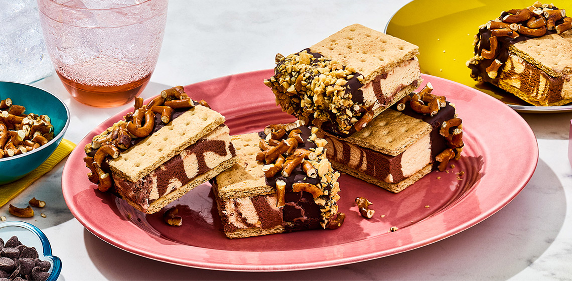 Four Peanut Butter S’mores Ice Cream Sandwiches on a plate with a small bowl of chopped pretzels off to the side and another plate with a single sandwich.