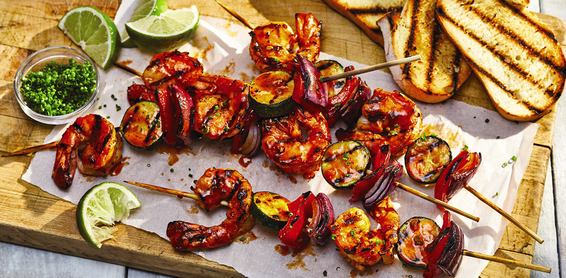 A wooden board with four skewers of Piri Piri Shrimp Skewers with bread, lime wedges and a small bowl of sauce.