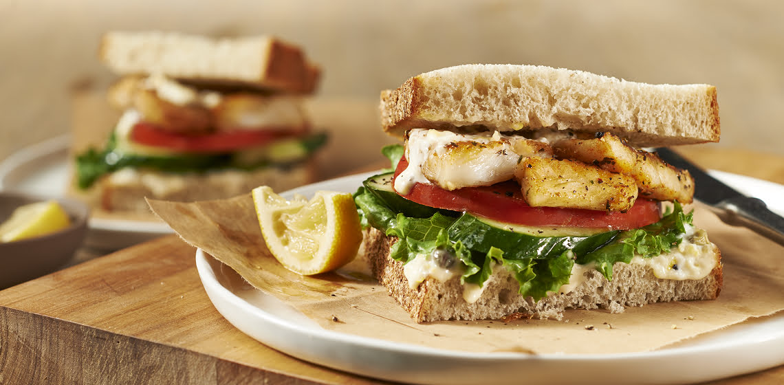 A Po’ Boy-Style Cod Sandwich on a plate with a knife and a lemon wedge sitting on a wooden cutting board with another sandwich and a bowl of lemon wedges in the background.