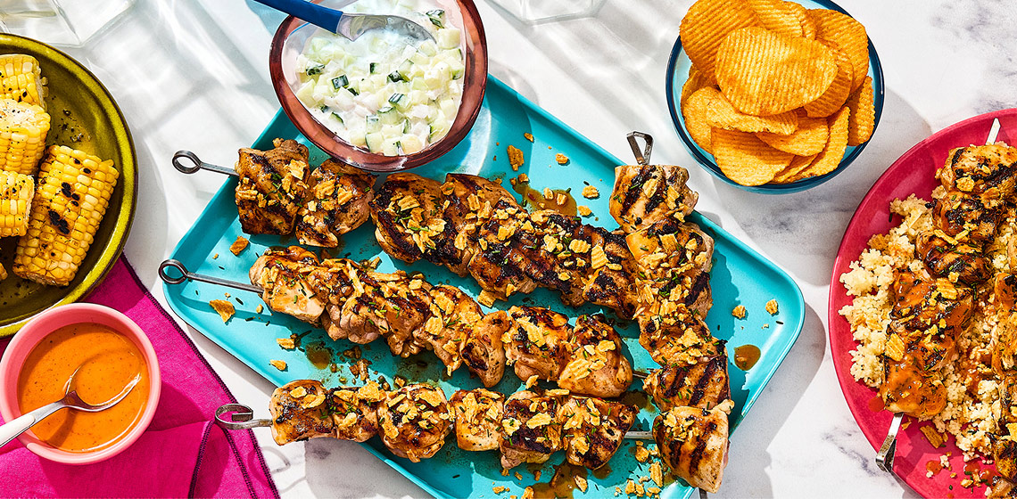 A platter of four Portuguese-Inspired Piri Piri Hot Honey BBQ Chicken Kabobs with a sauce, a small plate with more skewers, rice, and corn, and chips, corn, and more sauce in bowls off to the side.