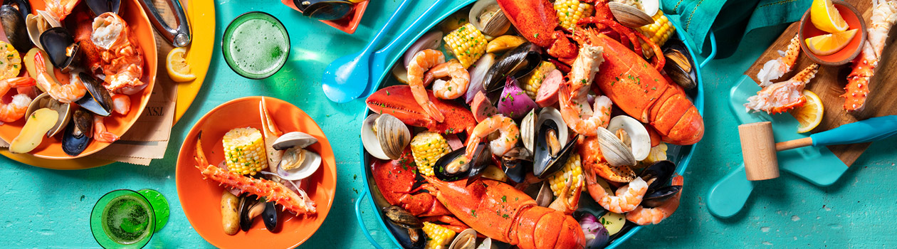 Brimming blue cast iron pot filled with shrimp, lobster claws, crab legs, potatoes and sausages with one bowl served up and drinks served.