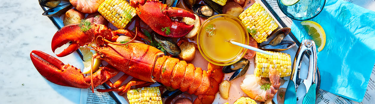 Large platter of a lobster boil with lobster, corn, shrimp, mussels, potatoes, sausage, and a bowl of sauce with a pair of small tongs.