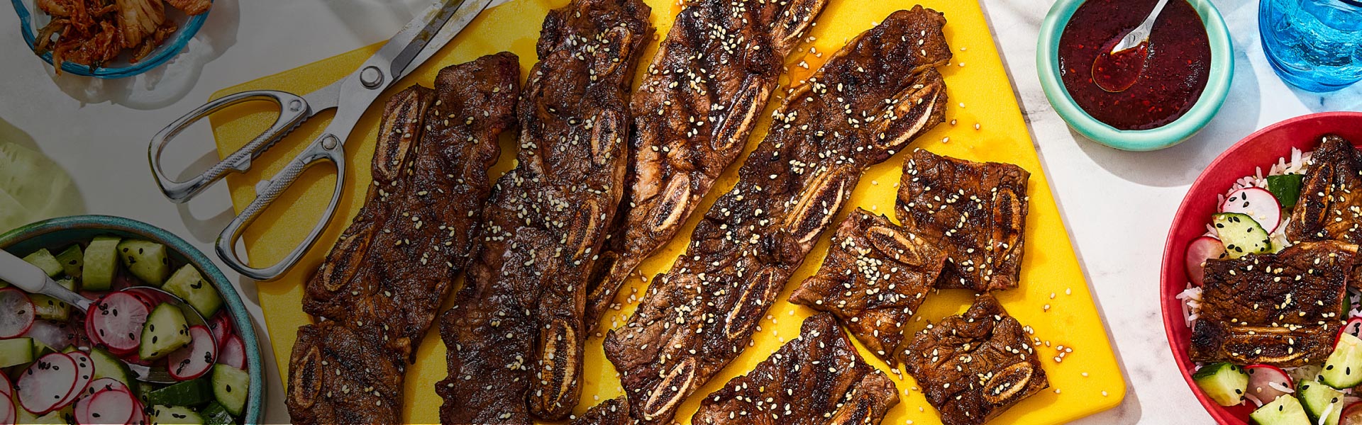 Overhead shot of kalbi Korean-style ribs on a yellow platter with scissors and a bowl of fresh salad on the side.