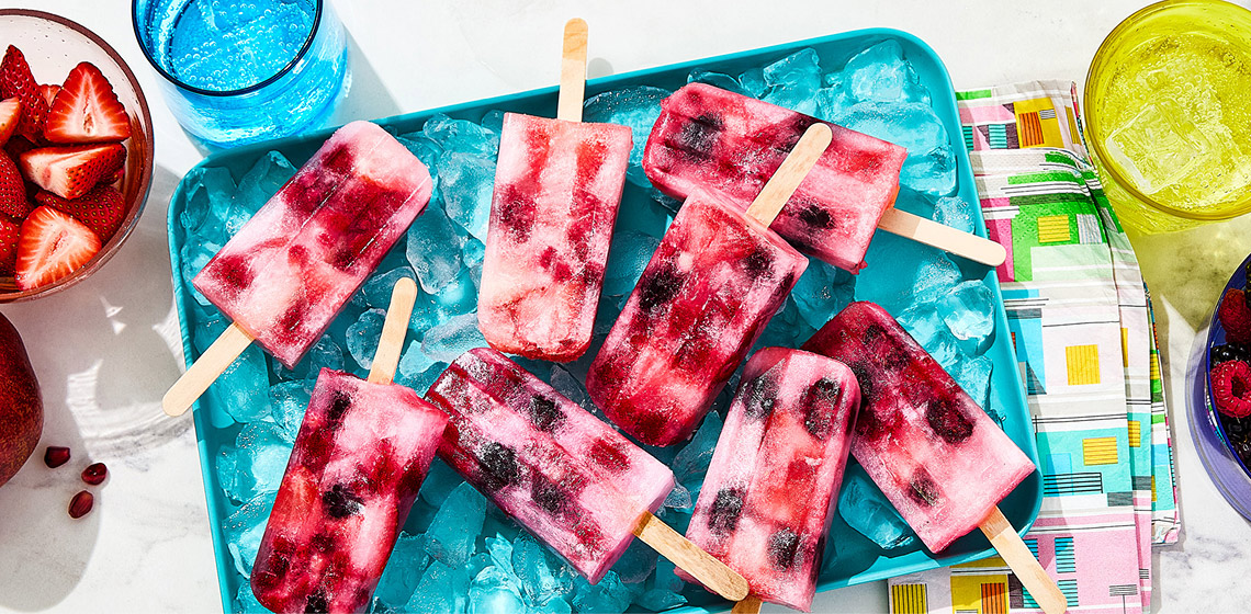 A platter filled with ice and several Sparkling Water Popsicles lying on it with bowls of berries and half a pomegranate off to the side.