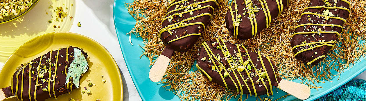 Four Dubai Inspired Chocolate Pistachio Bars on a bed of kataifi with one bar off to the side with a bite taken out and a bowl of pistachios.