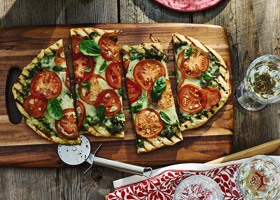 A cut up grilled tomato pizza on a wooden board with a pizza cutter also on the board and some filled glasses around it.
