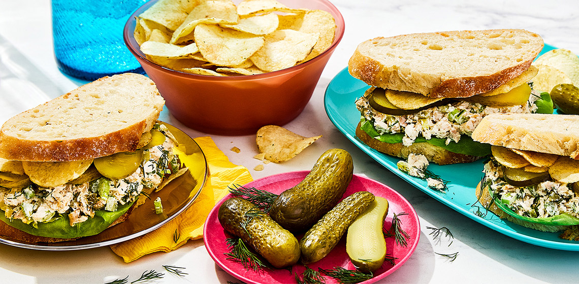 Three triple dill pickle chicken salad sandwiches, a plate of pickles and a bowl of chips.