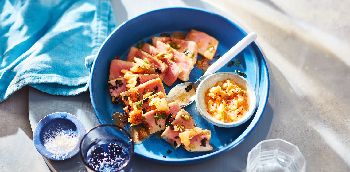 Slices of tuna steak and in a blue shallow bowl with a spoon and smaller bowl both filled with kimchi butter sauce.