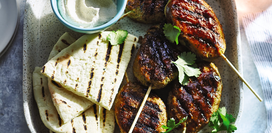 Five vegan chickpea kofta skewers, grilled naan slices and vegan sour cream on a grey plate.