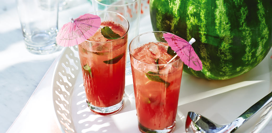 A white serving platter with watermelon rum punch in a hollowed-out watermelon and two tall glasses, and a silver serving ladle.