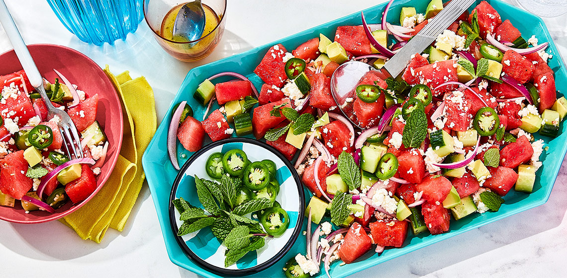 Watermelon salad on a teal platter with a bowl of jalapenos and mint with two bowls off to the side, one with sliced limes and one with more salad and a fork.