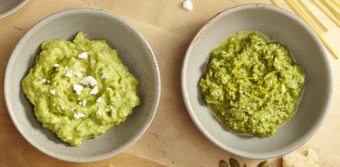 Pesto in a light grey bowl on a wooden board with cheese, pumpkin seeds and spinach leaves beside it.