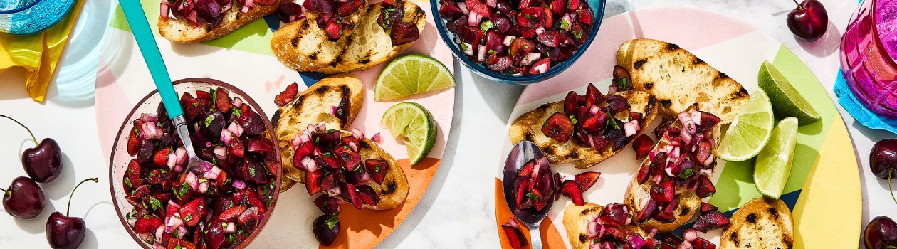 Two plates with crostinis topped with the cherry salsa and sliced limes, with bowls of the salsa and cherries on and around the plates.