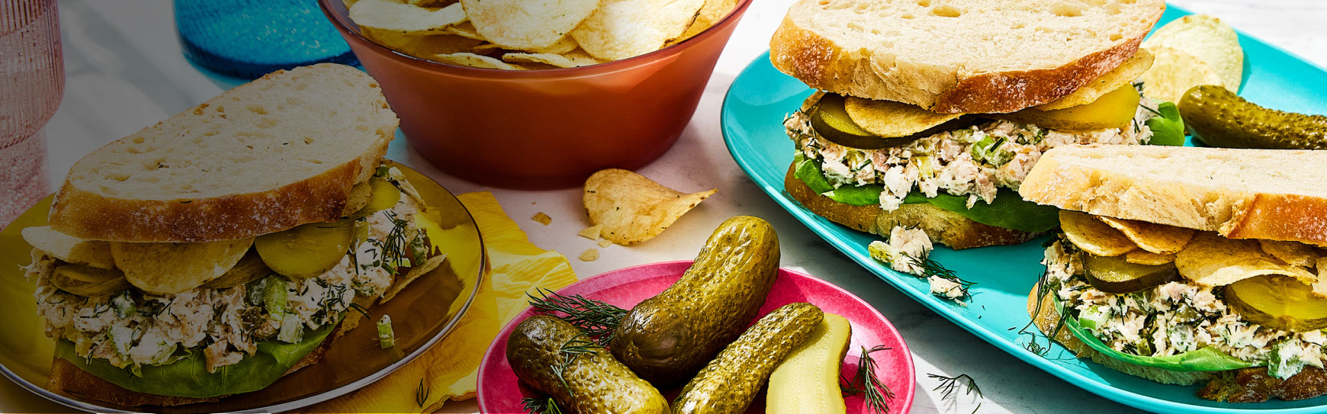 Three triple dill pickle chicken salad sandwiches, a plate of pickles and a bowl of chips.