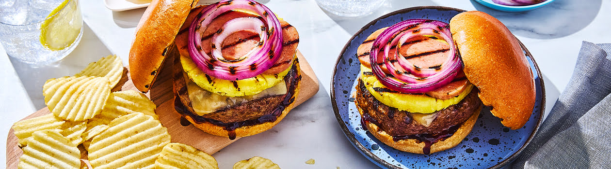 Two Hawaiian-inspired burgers on a white background with grilled pineapples and chips and two glasses of lemon water.