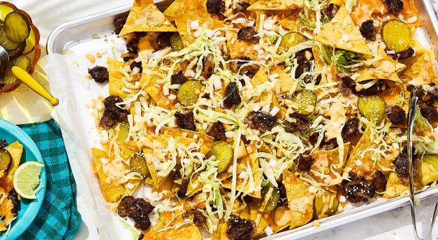Smashed burger nachos on a sheet pan and small plates, with small bowls of pickle slices and limes on the side.