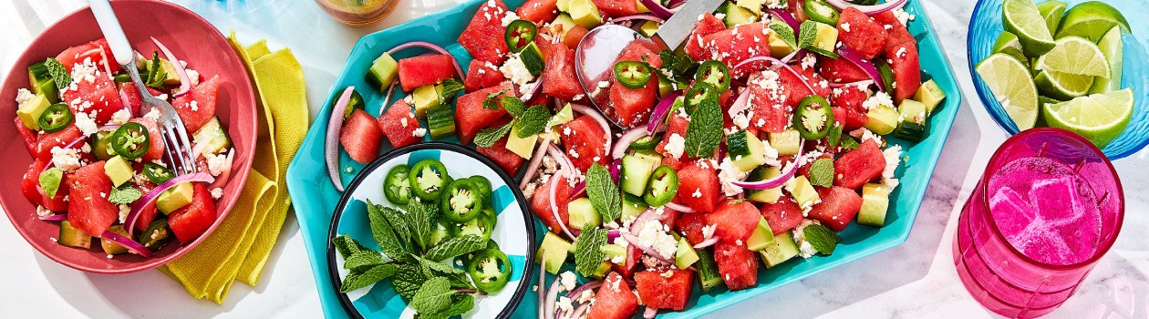 Watermelon salad on a teal platter with a bowl of jalapenos and mint with two bowls off to the side, one with sliced limes and one with more salad and a fork.