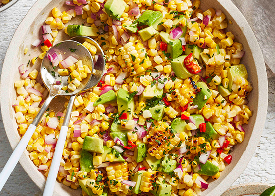 Overhead shot of Avocado & Corn Salad in a white serving bowl with two glasses of water and a small plate of cut limes next to it.