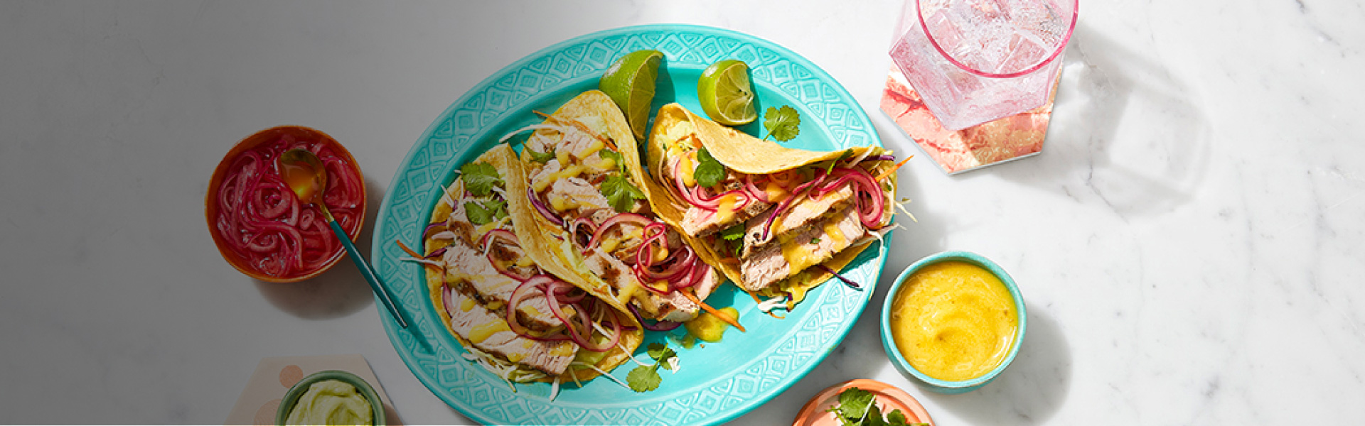 A teal plate with three grilled mahi mahi tacos and lime wedges with small bowls with toppings all around it.
