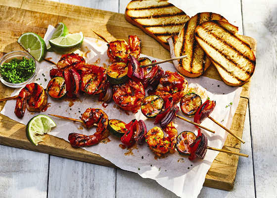 A wooden board with four skewers of Piri Piri Shrimp Skewers with bread, lime wedges and a small bowl of sauce.