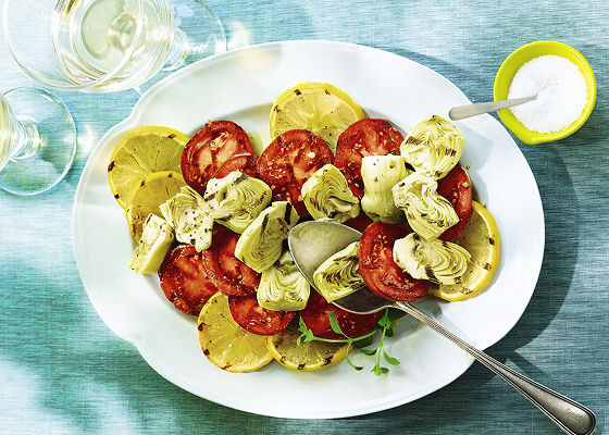 Grilled tomatoes, artichokes and lemons on a white plate with a spoon, and two wine glasses with white wine on a blue placemat