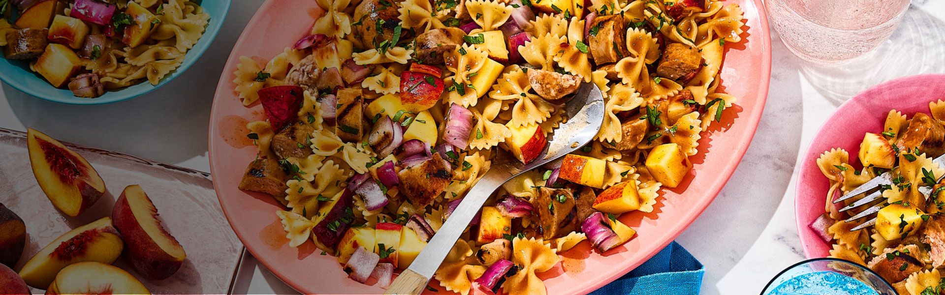 Platter filled with bowtie pasta salad and fresh peaches, with two bowls with the same pasta to the side.