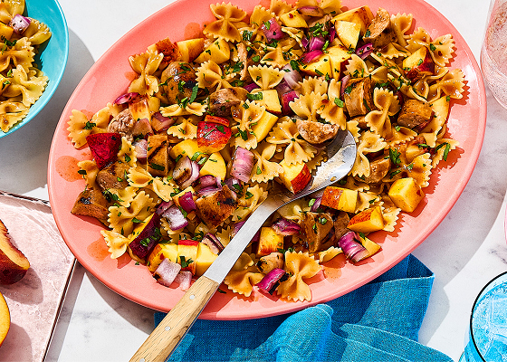 Platter filled with bowtie pasta salad and fresh peaches, with two bowls with the same pasta to the side.