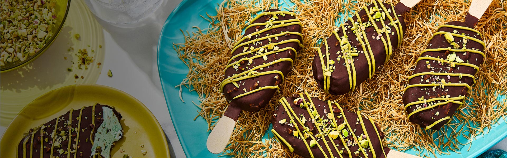 Four Dubai Inspired Chocolate Pistachio Bars on a bed of kataifi with one bar off to the side with a bite taken out and a bowl of pistachios.