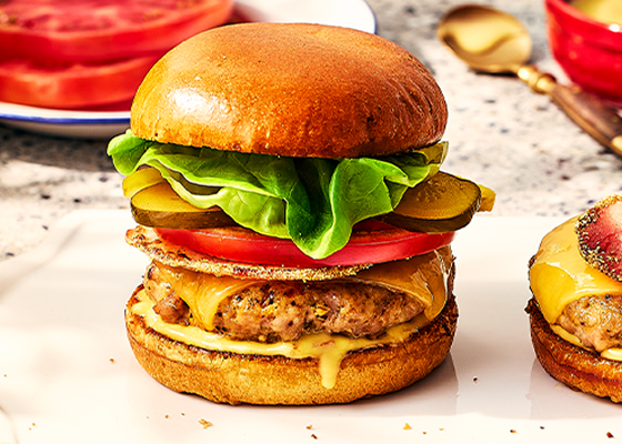 Pork and peameal bacon burger stacked high in a brioche bun, topped with pickles, tomato and cheddar cheese.